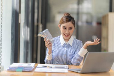 Genç Asyalı kadın masada oturuyor ve elinde Amerikan doları banknotuyla gülümsüyor ve başarılı bir iş fikri, maaş ve kâr kavramına bakıyor. Yardım almaktan mutluluk duyarım.