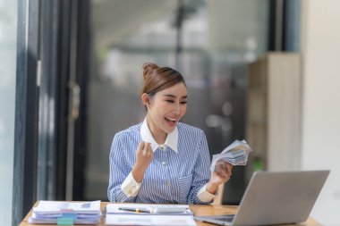 Genç Asyalı kadın masada oturuyor ve elinde Amerikan doları banknotuyla gülümsüyor ve başarılı bir iş fikri, maaş ve kâr kavramına bakıyor. Yardım almaktan mutluluk duyarım.