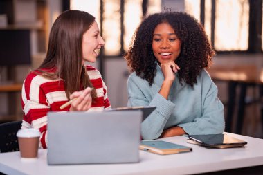 Fazla mesai yapan iki iş kadını ofiste sohbet ediyorlar. Dizüstü bilgisayarlarla, belgelerle, grafiklerle, ofisteki ışıklarla finansal analizlerle çalışıyorlar.