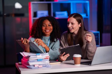 Fazla mesai yapan iki iş kadını ofiste sohbet ediyorlar. Dizüstü bilgisayarlarla, belgelerle, grafiklerle, ofisteki ışıklarla finansal analizlerle çalışıyorlar.