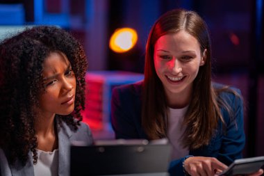 Fazla mesai yapan iki iş kadını ofiste sohbet ediyorlar. Dizüstü bilgisayarlarla, belgelerle, grafiklerle, ofisteki ışıklarla finansal analizlerle çalışıyorlar.