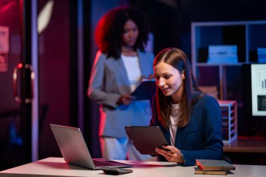 Fazla mesai yapan iki iş kadını ofiste sohbet ediyorlar. Dizüstü bilgisayarlarla, belgelerle, grafiklerle, ofisteki ışıklarla finansal analizlerle çalışıyorlar.