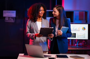 Fazla mesai yapan iki iş kadını ofiste sohbet ediyorlar. Dizüstü bilgisayarlarla, belgelerle, grafiklerle, ofisteki ışıklarla finansal analizlerle çalışıyorlar.