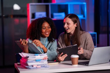 Beyaz iş adamı ve Afrikalı kadın takım olarak çalışır ve gece araştırmaları için laptop tableti kullanırlar. Veri analizi sonuçları dijital teknolojiyle karşılaştırma ve planlama