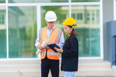 Asyalı erkek ve kadın mühendisler dış işleri sıkı şapkalar ve emlak acenteleri veya yöneticiler içinde inceleyerek dijital tabletlerle proje planlarını tartışıyor..