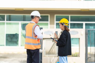 Asyalı erkek ve kadın mühendisler dış işleri sıkı şapkalar ve emlak acenteleri veya yöneticiler içinde inceleyerek dijital tabletlerle proje planlarını tartışıyor..
