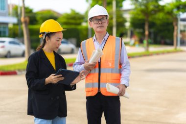 Asyalı erkek ve kadın mühendisler dış işleri sıkı şapkalar ve emlak acenteleri veya yöneticiler içinde inceleyerek dijital tabletlerle proje planlarını tartışıyor..
