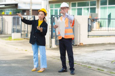 Asyalı erkek ve kadın mühendisler dış işleri sıkı şapkalar ve emlak acenteleri veya yöneticiler içinde inceleyerek dijital tabletlerle proje planlarını tartışıyor..