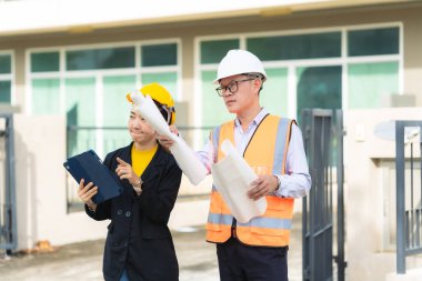 Asyalı erkek ve kadın mühendisler dış işleri sıkı şapkalar ve emlak acenteleri veya yöneticiler içinde inceleyerek dijital tabletlerle proje planlarını tartışıyor..