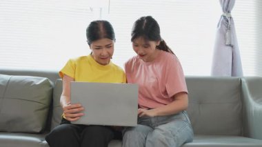 Asyalı anne ve kız kanepede dizüstü bilgisayarda birlikte oynuyorlar, internette bilgi arıyorlar, sosyal medyada geziniyorlar..