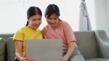 Asyalı anne ve kız kanepede dizüstü bilgisayarda birlikte oynuyorlar, internette bilgi arıyorlar, sosyal medyada geziniyorlar..
