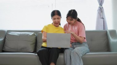 Asyalı anne ve kız kanepede dizüstü bilgisayarda birlikte oynuyorlar, internette bilgi arıyorlar, sosyal medyada geziniyorlar..