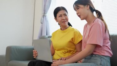 Asyalı anne ve kız kanepede dizüstü bilgisayarda birlikte oynuyorlar, internette bilgi arıyorlar, sosyal medyada geziniyorlar..