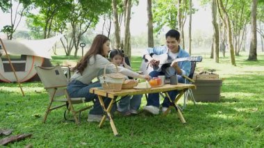 Mutlu Asyalı aile parkta kamp yapıyor, dışarıda eğleniyor, bir tatil partisini kutluyor, yemek yiyor, gitar çalıyor ve birlikte şarkı söylüyor..