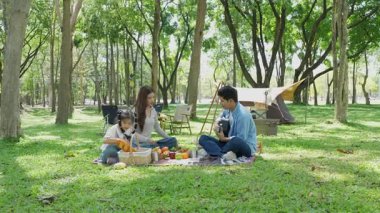 Mutlu Asyalı aile parkta kamp yapıyor, dışarıda eğleniyor, bir tatil partisini kutluyor, yemek yiyor, gitar çalıyor ve birlikte şarkı söylüyor..