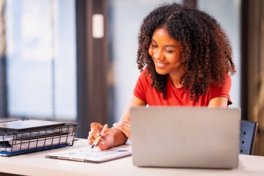Afro-Amerikalı genç kız geceleri dizüstü bilgisayar ve kitaplarla ders çalışıyor ve oturma odasındaki masada oturan dizüstü bilgisayar öğrencilerini kullanarak işe odaklanıyor.