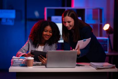 İki kendine güvenen iş kadını, gece geç saatlerde modern ofiste şirket projeleri üzerinde çalışan raporlarla dosyaların paylaşıldığı bir toplantıda dizüstü bilgisayar kullanırken strateji planlıyorlar..