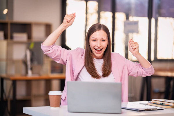 Молодая женщина радостно празднует успех за столом с ноутбуком. Поднимите руки с радостью Волнение и финансовый успех