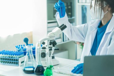 Laboratuar laboratuarında mikroskop deneyi sırasında plastik şişeden deney kabına mavi sıvı kimyasal döken laboratuvar önlüklü kadın doktor..