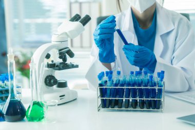 Laboratuar laboratuarında mikroskop deneyi sırasında plastik şişeden deney kabına mavi sıvı kimyasal döken laboratuvar önlüklü kadın doktor..