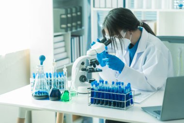 Laboratuar laboratuarında mikroskop deneyi sırasında plastik şişeden deney kabına mavi sıvı kimyasal döken laboratuvar önlüklü kadın doktor..