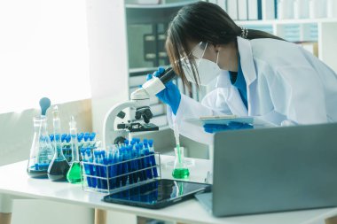 Laboratuar laboratuarında mikroskop deneyi sırasında plastik şişeden deney kabına mavi sıvı kimyasal döken laboratuvar önlüklü kadın doktor..