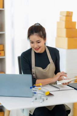 Genç Asyalı kız evde kendi işi olan bir serbest çalışan. Dizüstü bilgisayarla çalışan genç kadın, KOBİ işine çevrimiçi satış, pazarlama, paketleme, teslimat, KOBİ ve e-ticaret konseptiyle başladı.