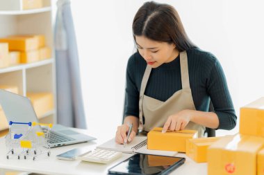 Genç Asyalı kız evde kendi işi olan bir serbest çalışan. Dizüstü bilgisayarla çalışan genç kadın, KOBİ işine çevrimiçi satış, pazarlama, paketleme, teslimat, KOBİ ve e-ticaret konseptiyle başladı.