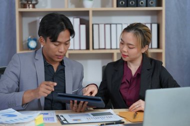 Resmi giyinmiş iki Asyalı iş adamı bir masada birlikte çalışıyor, gülümsüyor ve iş hakkında konuşuyorlar. Finansal tabloyu göster ve bir tablet ve dizüstü bilgisayarla bir ofis masasında işbirliği yap.