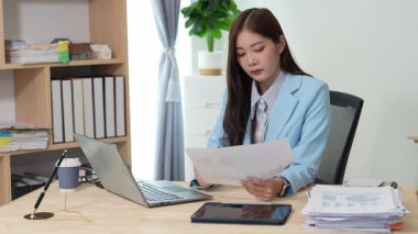 Bilgisayarı ve finansal belgeleri olan genç Asyalı kadın çalışanlar ofis masasında gülümsüyor ve grafikleri gösteren bilgisayarlarla çalışıyor. başlangıç iş fikri