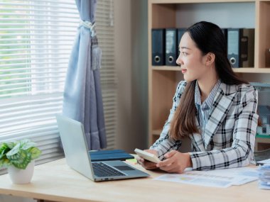 Asyalı iş kadını kahve içiyor ve ofis masasındaki hesap makinesi ve dizüstü bilgisayarla ofiste mali belgeler üzerinde çalışıyor..