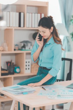 Asyalı iş kadını, masada grafik dökümanı ve dizüstü bilgisayarı olan müşteriyle konuşmak için akıllı telefon kullanıyor. Kısa fikirlerle çevrimiçi kadın toplantısı. 