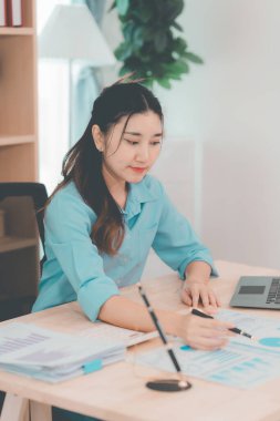 Genç Asyalı muhasebeci masada oturmuş belgeleri analiz ediyor. Şirket projeleri üzerinde çalışan grafikleri analiz ediyor..