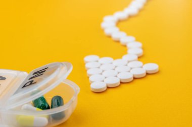 Ok şeklinde beyaz tıbbi haplar plastik günlük düzenleyiciyi (bir haftalık ilaç) işaret eder ve ona doğru akar. Sarı arkaplanda yakın plan.
