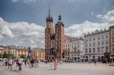 10 Temmuz 2024. Turistler Polonya 'nın Krakow kentindeki St. Marys Bazilikası ile tarihi Ana Pazar Meydanı' nı parçalı bulutlu bir gökyüzü altında keşfediyorlar.