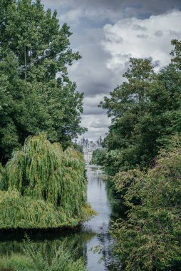 Londra 'nın tarihi sarayının manzarası yemyeşil ağaçların arasından görülüyor, sakin bir nehir ve bulutlu bir gökyüzünün altında köprüsü var.