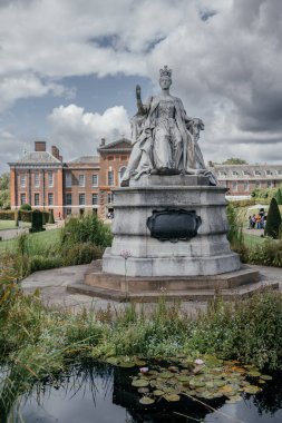 Kensington Sarayı 'nın bahçesindeki Kraliçe Victoria heykeli. Arkasında tarihi kırmızı tuğla sarayı olan parçalı bulutlu bir günde.