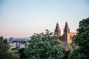 Şehir çatıları ve günbatımında yeşil ağaçlarla kaplı Kelvingrove Sanat Galerisi ve Müzesi 'nin uzak manzarası Glasgow kültürel ve mimari mirasını vurguluyor.