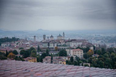 Bergamo, İtalya 'nın panoramik manzarası, tarihi mimari, kırmızı çatılar, kilise kuleleri ve bulutlu bir gökyüzünün altındaki tepeleri gösteriyor.
