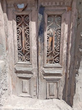 Bu görüntü, her biri karmaşık demir işçiliği ve desenlerle süslenmiş bir çift yıpranmış ahşap kapının kırsal cazibesini yakalar. Kapılar ustalığın bir göstergesi olarak duruyor. Yıllanmış odunları, geçmiş yılların hikâyelerini anlatıyor. Soyulmuş boyaya rağmen 