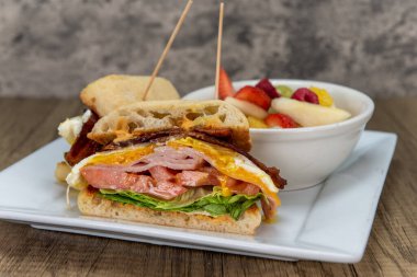 Taze pişmiş ciabatta rulosu bir kase taze meyveyle servis edilen devasa bir kahvaltı kulübü sandviçi içeriyor..