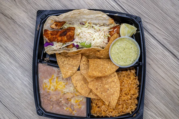Overhead view of to go order of halibut fish taco meal with beans and chips all served in plastic container with dressing on the side.