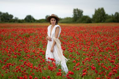 Beyaz elbiseli ve şapkalı genç bir kadın yaz gelinciklerinin arasında poz veriyor.