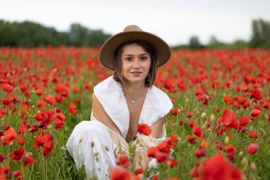 Beyaz elbiseli ve şapkalı genç bir kadın yaz gelinciklerinin arasında poz veriyor.