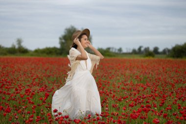Beyaz elbiseli ve şapkalı genç bir kadın yaz gelinciklerinin arasında yürüyor.