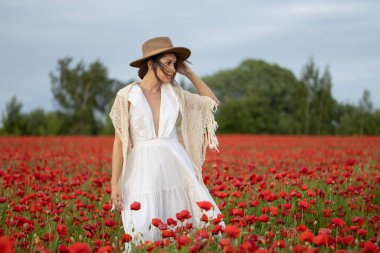 Beyaz elbiseli ve şapkalı genç bir kadın yaz gelinciklerinin arasında poz veriyor.
