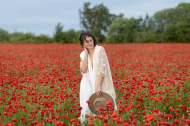 Beyaz elbiseli ve şapkalı genç bir kadın yaz gelinciklerinin arasında poz veriyor.