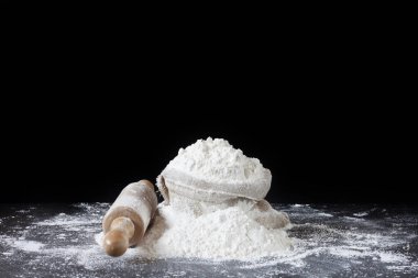 Masaya yayılmış beyaz toz ve silindirli buğday unu torbası siyah arka planda.