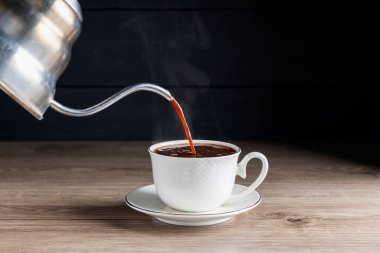 Koyu siyah arka planda, ahşap bir masada kavrulmuş kahve çekirdekleri ile beyaz bir kahve bardağına sıcak kahve dökmek..