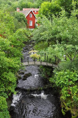 Yeşil yapraklar ve arka planda kırmızı ahşap binalarla çevrili kemer şeklinde yaya köprüsü olan güzel bir dere..